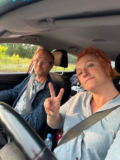 This is a candid, portrait-oriented photograph taken from inside a car, likely on a sunny day given the bright lighting. The camera is tilted, suggesting it was held by one of the passengers.
In the foreground, a woman with vibrant red, curly hair is looking towards the camera. She is wearing a light blue, textured shirt and a seatbelt. Her facial expression appears relaxed, with a slight smile. Her eyes are blue and she has freckles.
Behind her and slightly to the right, a man is visible, also wearing a seatbelt. He has short, light brown hair and is wearing glasses and a dark blue patterned shirt over a lighter patterned t-shirt. He is making a peace sign with his right hand, and his expression is also relaxed.
The background of the photograph shows a glimpse of the exterior through the car windows. Green trees and a fence are visible, suggesting a rural or semi-rural setting. The light suggests it is either morning or late afternoon.
There is a water bottle with a blue cap visible on the passenger side of the car. Some luggage or bags are also present in the back seat. The interior of the car, including the car's sun visor and interior panels, are also visible.
The overall mood of the photograph is casual and suggests a road trip or outing with a couple. There is no visible text in the image.