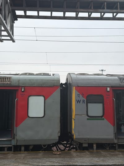 This is a close-up, eye-level, medium shot of two adjacent train carriages at the Ballabgarh railway station in India. The carriages are primarily red and gray with diagonal red markings on the left carriage. The right carriage has "प रे WR" and "EXIT" printed on it. Both carriages have windows and doors. Overhead, power lines and a metal structure are visible against a cloudy sky. The train is positioned on tracks beside a platform.