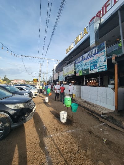 This image captures a vibrant street scene in Kimorori, Kenya, under a clear, bright blue sky, suggesting either morning or late afternoon due to the prominent shadows cast by the sun.
The scene depicts a commercial strip with a row of single-story buildings housing various businesses on the left. A large blue sign prominently displays "SAN'S BUTCHERY," indicating a meat shop. Below this, the word "MPESA" is visible, signifying the presence of a popular mobile money transfer service in Kenya. Other signs are present along the shopfronts, including what appears to be "AFRICAN PLAZA." Overhead, numerous power lines or communication cables crisscross the sky, and a string of decorative lights is strung across the street, possibly for illumination or festivities.
The ground is unpaved, consisting of dirt or gravel. Along this path, several colorful plastic containers and buckets (green, white, orange) are lined up, some supported by sticks or poles. These containers might be part of market stalls or used by vendors for displaying goods. Several people are visible in the mid-ground, standing near these containers and the shopfronts. One individual in a white top and red apron is notably positioned near a green and white container, possibly a vendor or shopkeeper.
On the right side of the street, multiple cars are parked, indicating a parking area adjacent to the commercial activity. The overall impression is that of an active, everyday commercial and market environment.