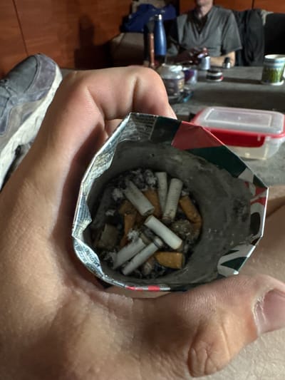 A close-up, overhead shot shows a person's hand holding an open, crumpled cigarette pack filled with numerous cigarette butts. The cigarette butts are of varying colors, some white and some with brown filters. Ash is also visible within the pack. The pack itself has a colorful design with green and red accents, and some text is visible along the inner rim. The person's thumb and fingers are visible in the foreground, and part of their leg and foot, wearing a grey shoe, are visible in the lower portion of the image. In the background, out of focus, are other objects including a red and white plastic container, some bottles, and what appears to be fishing equipment. The scene appears to be indoors or in a sheltered outdoor area given the lighting and background elements. There are no clear indications of the time of day or weather. The overall impression is one of casual disposal of smoking waste. The text on the cigarette pack is partially visible, featuring Polish words like "PUSZCZOWE" and "NASYC".