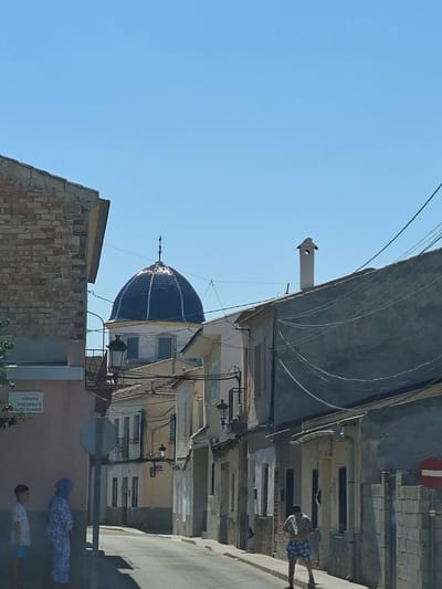 The image depicts a street scene in Granja De Rocamora, Spain, dominated by a bright blue domed building visible in the background. The street is lined with buildings of varying architectural styles and states of repair. On the left side, a brick building is partially visible. In the foreground, two individuals are standing on the sidewalk next to a street sign. One is a child wearing a white t-shirt and blue shorts, and the other is a person wearing a blue head covering and a patterned blue and white outfit. Further down the street, another person is standing, wearing a patterned shirt and blue shorts. Overhead, power lines crisscross the sky. A red and white circular sign indicating a prohibition is visible on the right side of the street. The sky is clear and blue, suggesting a sunny day. The overall impression is of a quiet, sun-drenched street in a Spanish town.