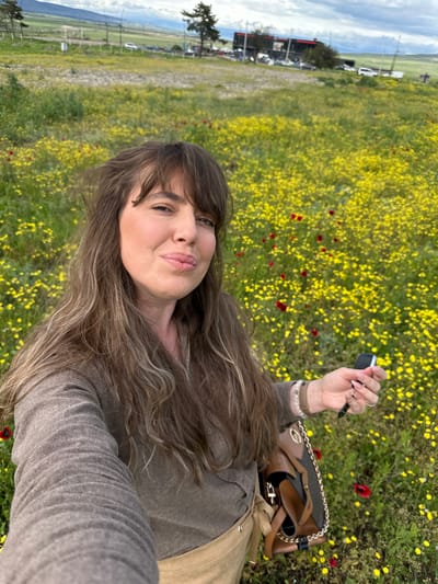 The image shows a woman in a field of yellow flowers with some red flowers. She is holding a car key. In the background, a road with parked cars can be seen, as well as mountains and the sky. Marneuli is not apparent in the image.