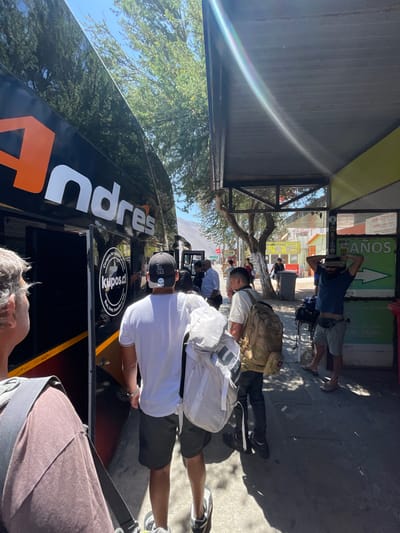People wait to board an "Andres" bus on a sunny day. A man wearing a hat and taking pictures stands near a "BAÑOS" sign. Some passengers have backpacks.  A black baseball cap with a logo is visible on one passenger.  The bus has the address "Kupos.cl" printed on it. The scene suggests a bus station or transport hub with people preparing for a journey.