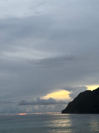 This is a photograph of the sea and a mountainous coastline at dusk. The sky is filled with dark, heavy clouds, with a bright yellow glow near the horizon where the sun is setting behind the mountain. The sea is a deep gray-blue, with gentle waves reflecting the light from the sky. A dark, rugged mountain juts out from the right side of the frame, its silhouette stark against the colorful sky. The location is Meunasahbale, Indonesia.