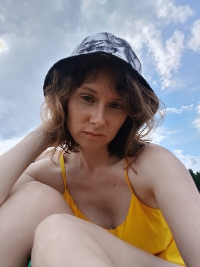 A woman with shoulder-length brown hair and freckles is wearing a bucket hat with a palm tree pattern and a yellow swimsuit. She is looking at the camera with a neutral expression, her left arm bent and her hand resting on her head. The background is a cloudy blue sky, with trees visible on the right side. The photograph was taken outdoors, possibly near a body of water, given the relaxed attire and the natural setting. It is not possible to determine the specific location in Revda, Russia, from the image alone.