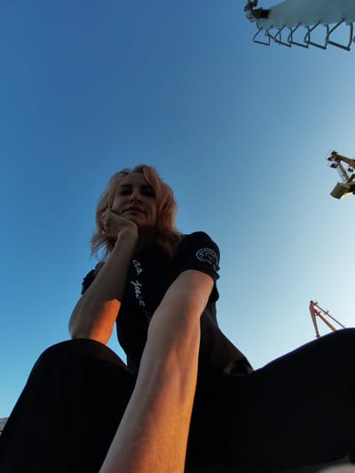 A low-angle, wide shot captures a woman with blonde, shoulder-length hair sitting outdoors against a clear blue sky. She is wearing a black t-shirt and black pants, and her left arm is bent with her hand resting on her chin. A ring is visible on her left index finger. The sunlight casts a warm glow on her arm and cheek. In the background, the tops of two industrial cranes are visible against the sky, suggesting an industrial or port area. The lower portion of a third structure, appearing to be a crane or similar industrial equipment, is visible in the upper left of the frame. The overall impression is that of a portrait taken at dusk or dawn in an outdoor industrial setting.