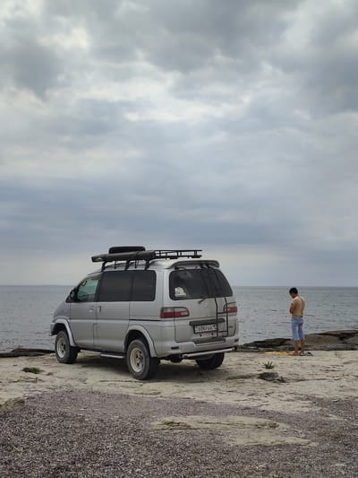 A silver off-road van is parked on a rocky shore near the Caspian Sea in Aqshuqyr, Kazakhstan. A young man, wearing a light brown t-shirt and blue shorts, stands facing away from the van, looking down at a yellow object on the ground. The van is equipped with a roof rack and a spare tire mounted on the back. The sky is overcast with gray clouds. The ground is covered in small pebbles and rocks.