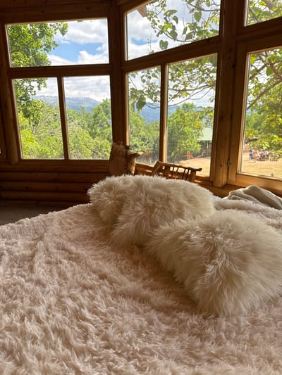 A view from inside a log cabin in Gornje Mrke, Montenegro, features a large multi-pane window showing a lush green mountainous landscape with trees and a glimpse of a grassy field in the distance with some people sitting. Inside, a white shaggy blanket and two round, fluffy pillows are visible. A wooden chair sits beside the window. The interior walls are constructed from logs. The sky is blue with scattered clouds.