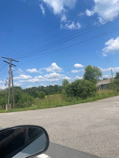 The image depicts a rural roadside scene in the vicinity of Pomogalovo, Russia, under a clear blue sky with scattered white clouds. A gray asphalt road curves slightly through the frame. On the left side of the road, a wooden utility pole with multiple wires is visible, alongside overgrown green vegetation and trees. In the background, a dense line of trees forms a natural boundary, and further to the right, an aged, weathered wooden house with a gray roof stands among the foliage. A side-view mirror of a vehicle is partially visible in the lower left corner, reflecting a person's arm and hand. The overall impression is of a quiet, possibly abandoned, rural setting on a bright, sunny day.
