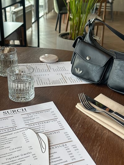 This image captures a close-up, overhead view of a wooden dining table inside "SURCH COFFEE & KITCHEN" located in Усады, Republic of Tatarstan, Russia.
On the left side of the table, two clear, fluted glass tumblers filled with water are positioned. To their right, a small, white, oval-shaped object with faint illegible text rests on the table surface.
Two white paper menus are displayed. One menu, largely visible in the lower-left to middle-bottom portion of the frame, features "SURCH COFFEE & KITCHEN" at the top and lists various categories such as "КОФЕ" (Coffee), "ЛИСТОВОЙ ЧАЙ" (Loose Leaf Tea), "ФИРМЕННЫЙ ЧАЙ" (Signature Tea), "МОЛОКОМ" (with Milk), "КОКТЕЙЛИ" (Cocktails), "Салаты" (Salads), "Горячие блюда" (Hot Dishes), and "ЛИМОНАДЫ" (Lemonades), with corresponding prices in Rubles and volume measurements in milliliters. A second menu is partially visible further back, behind the glasses and a bag.
Dominating the right side of the image is a black faux-leather shoulder bag featuring prominent Adidas branding. The bag has two flap pockets; one on the front features a silver Adidas Trefoil logo, and a smaller one above it has a silver metallic button. A silver Adidas Trefoil logo charm or pull is attached to the zipper closure. The bag's black strap and a silver buckle are also visible.
On the far right, a silver fork and knife with dark handles are neatly arranged on a folded light beige fabric napkin. The knife blade shows "Dayinci" engraved.
The background reveals a modern cafe interior with blurred elements including light-colored wooden tables, dark-framed chairs with light upholstery, large windows, dark metal room dividers, a potted green plant, and light-colored flooring, indicating an active dining establishment. No individuals are present in the frame.