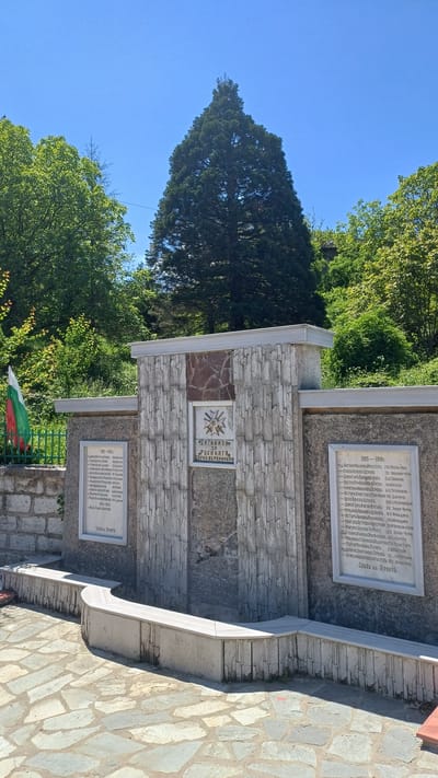 The image shows a stone monument in Gorna Verenitsa, Bulgaria. The monument is made of light gray stone blocks and contains three plaques with text. The top plaque lists names from 1915-1918, and the bottom plaque lists names from 1941-1945. The central plaque features an emblem and the words "Загинали за родината Горна Вереница" (Fallen for the homeland Gorna Verenitsa). The Bulgarian flag is visible next to the monument. Behind the monument are trees and a blue sky. The monument is located next to a stone-paved area.