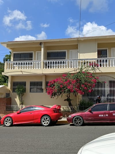 The image depicts a street scene in Puerto Plata, Dominican Republic, on a bright, sunny day with scattered white clouds in a vibrant blue sky. The focus is on a light yellow, multi-story building with white railings and decorative white window grates. Bougainvillea with vibrant pink flowers cascades from the upper balcony, adding a splash of color to the architecture.
Parked along the street are two cars. A maroon sedan is parked in front of the building, and a bright red sports car is parked in the foreground, facing away from the viewer. The red car has a distinctive spoiler and prominent multi-spoke wheels. The maroon car has what appears to be Japanese text written vertically on its side.
The setting is residential, with trees and foliage visible, suggesting a warm climate. The lighting indicates it is daytime, likely mid-morning or afternoon. The overall impression is of a peaceful, everyday scene in a tropical or subtropical urban environment.