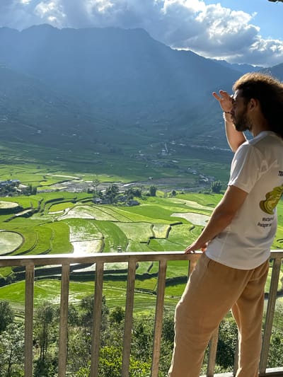An adult male with long, curly dark hair and a beard stands on a balcony, viewed from the rear-right, overlooking a vast landscape in Tú Lệ, Vietnam. He wears a white short-sleeved t-shirt with a light green abstract design and partial text "WHATSA..." and "Instagram: m..." on the back, along with light brown long pants. His right hand rests on a grey metal railing, while his left hand is raised to shield his eyes, indicating he is looking toward the left. The background features a wide valley filled with numerous vibrant green rice terraces, some containing standing water that reflects sunlight. A light-colored river or stream winds through the valley floor, bordered by scattered small residential buildings and trees. Layers of distant mountains, appearing blueish due to atmospheric haze, rise under a partly cloudy sky with patches of bright blue. The scene is brightly illuminated by natural sunlight.