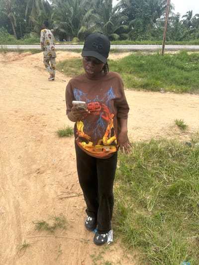 The image shows a young woman standing on a dirt path, looking at her cell phone. She is wearing a brown sweatshirt with a graphic design, black pants, and black Crocs. She has on a black baseball cap. In the background, another person in a patterned outfit walks away from the camera. The setting appears to be rural, with palm trees and other vegetation visible. A paved road runs in the distance. The location is identified as Ganyingbo, Nigeria.