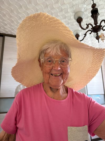An elderly woman wearing a large straw hat and round gold-rimmed glasses smiles at the camera. She is wearing a pink shirt with a grey pocket. A chandelier is visible in the background. The location is Valozhyn, Belarus.