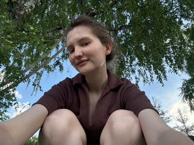 A young woman with light brown hair pulled back is sitting outdoors, looking up and towards the camera with a slight smile. She is wearing a dark brown, collared, v-neck knit top. Her legs are bent, with her knees close to her chest. The background consists of a blue sky with scattered white clouds, framed by the green leaves and branches of a large tree. The angle of the shot is from a low perspective, looking up at the woman and the tree canopy. The environment appears to be a natural outdoor setting, possibly a park or wooded area, in or near Burdyata, Russia.
