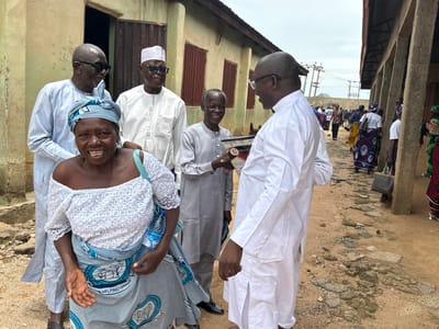In Kufana, Nigeria, a group of individuals are gathered outdoors near a weathered, yellow building with a corrugated metal roof. The foreground features a woman wearing a white, off-the-shoulder top and a blue and white patterned skirt, smiling as she holds a bottle of water. She stands beside a man in a light blue traditional outfit and sunglasses. To their right, a man in a white cap and traditional attire is speaking with another man in a gray outfit, who is holding a book. Further in the background, a crowd of people, many in colorful African prints, are visible, suggesting an event or gathering. The ground is a mix of dirt and broken pavement. The overall scene depicts a community interaction in a rural Nigerian setting.