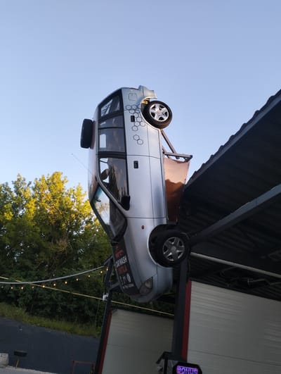 A silver car is mounted upside down on a metal structure, appearing to be a roadside attraction or advertisement. The car is positioned at an angle, with its wheels visible. Some geometric patterns are visible on the car's body. Part of a covered structure with a corrugated metal roof is visible to the right of the car. In the background, lush green trees are illuminated by sunlight, suggesting it's daytime. A string of lights is strung between poles in the middle ground. A sign on the car reads "CAR WASH UNTILA". Below the car, a digital display shows the word "BONUS" in purple letters. The scene appears to be outdoors, possibly in a rural or suburban area. The sky is clear and blue.