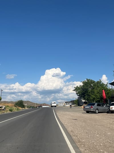 This is a medium shot of a roadside scene in Gladzor, Armenia. A paved road curves into the distance, bordered by a gravel shoulder on the left and dry grass and low hills on the right. A white line marks the edge of the road. Under a bright blue sky with large, puffy white clouds, a few vehicles are parked or driving. One is a grey sedan, and another appears to be a white van. A large green tree with full foliage stands near the parked cars, and a red umbrella is visible beneath its branches. In the distance, the landscape is dotted with sparse vegetation and rocky hills. The time of day appears to be midday, indicated by the bright sunlight and clear sky. The weather is warm and sunny. A few people are visible near the parked cars, suggesting a stop or a small gathering. No discernible emotions are conveyed. The license plate on the grey car shows the text "B57 GD 723".