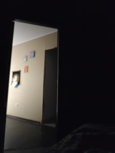 The image depicts an interior hallway, partially illuminated, with a doorway on the left offering a view into a room. On the wall of the room, there are three small square pictures. The leftmost picture is a dark banner with a portrait of a person in the center, featuring red and blue accents. To the right of the banner are two smaller square pictures, one orange and one light blue, each with an indistinct image inside. Below these pictures, a light switch is visible on the wall. The floor of the hallway is dark and reflective, suggesting a polished surface. The overall impression is of a dimly lit, private indoor space, possibly a residential hallway. The location of Шевели, Russia is not discernible from the visual information provided in the image.