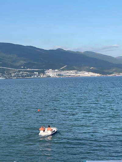 The image shows a bright, clear day with a vast expanse of blue sky and calm, rippling blue water. In the foreground, two people are relaxing on a white boat, which appears to be a small pedal boat. They are both wearing swim trunks and are seated facing forward. The boat is on the water, and a small orange buoy is visible nearby.

In the background, a coastline is visible, characterized by a densely populated area with buildings and roads. Beyond the coastal town, dark green mountains rise up, suggesting a coastal mountain range. The overall scene conveys a sense of leisure and relaxation on a sunny day at the seaside, with the backdrop of a developed coastal area and natural mountain scenery.

The lighting suggests it is daytime, likely late morning or afternoon, with the sun casting a bright glow on the water. No text is visible in the image. The setting appears to be a popular tourist or recreational spot where people can enjoy activities on the water. The location is identified as Chernikov, Russia.