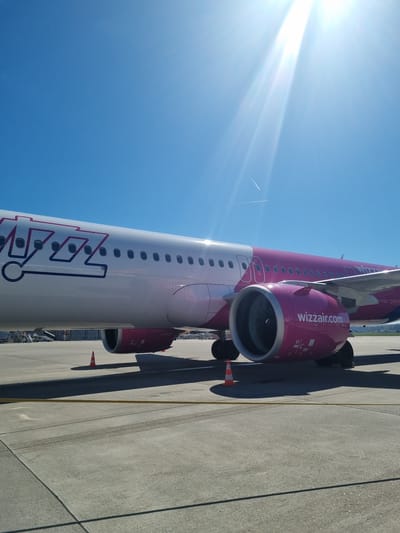 The image depicts a Wizz Air Airbus A321 aircraft parked on an airport tarmac. The fuselage is primarily white with pink and purple branding. Visible are the aircraft's engines, painted pink, with the "wizzair.com" logo displayed. The sky is blue, with the sun positioned in the upper left corner. A contrail from another aircraft is visible in the sky. The tarmac is concrete, marked with yellow lines, and includes an orange traffic cone. The location is the EuroAirport Basel-Mulhouse-Freiburg, which is located near Saint-Louis, France.