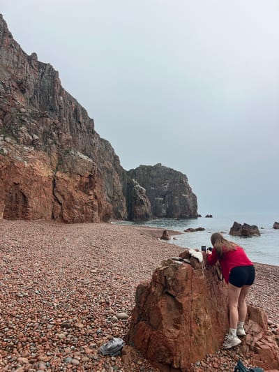 The image depicts a rocky beach in Russkiy, Russia. A woman wearing a red shirt and black shorts is leaning over a large rock near the water's edge, seemingly using a phone to take a picture. The beach is covered in small, reddish-brown pebbles. To the left, a steep, rocky cliff rises up. The sky is overcast, creating a muted color palette.