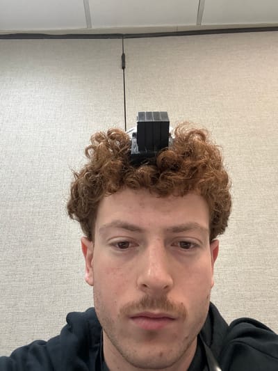 A close-up, frontal image depicts a young adult male with curly, reddish-brown hair and light skin. A black tefillin, specifically the *shel rosh* (head-tefillin) component consisting of stacked rectangular blocks with visible wiring, rests on top of his head, embedded in his hair. His brown eyes are directed downwards, and he has a light mustache. He wears a dark-colored hooded garment, with a dark lanyard or strap visible near his neck. The background features a light-colored, textured wall, likely an office partition, which meets a drop ceiling composed of light-colored acoustical tiles and a visible grid. A thin, black vertical wire is visible against the wall in the upper left portion of the frame. The image depicts an indoor environment. No specific visual identifiers within the image allow for the determination of its precise location within Fairfield, United States of America.