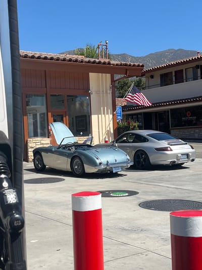 The scene depicts a sunlit outdoor forecourt in Montecito, United States. Two luxury automobiles are parked on a paved surface featuring circular utility covers and three red-and-white topped bollards in the immediate foreground. On the left, a light blue and silver two-tone vintage Austin-Healey 3000 convertible is parked with its hood open, revealing engine components; its soft top is lowered. To its right and slightly behind, a silver Porsche 911 coupe, with "Carrera" visible on its rear, is parked.

Behind the vehicles are two Mediterranean-style buildings, both featuring terracotta tiled roofs. The left structure has light brown wooden siding, large glass storefronts, and a visible surveillance camera. The right structure, also with a terracotta roof, displays an American flag and a partially visible sign for "STERLING HUDDLESON ARCHITECTURE." A black gas pump nozzle is partially visible on the far left. Mountains rise in the distant background under a clear blue sky. No individuals are present in the frame.