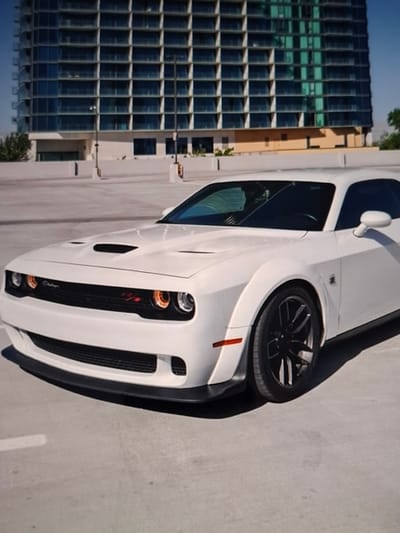 The image shows a white Dodge Challenger parked in an outdoor parking lot. In the background, a modern, multi-story building with a glass facade dominates the scene. The sky is clear and blue. The car's front end, including the grille, headlights, and hood scoop, is prominently featured. The parking lot surface appears to be concrete, with some faint white lines visible. There are no people or animals in the image. This scene does not depict the city of Urus-Martan, Russia.