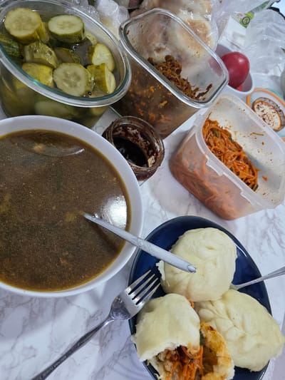 A close-up, overhead shot shows a table laden with Korean dishes, likely in Eumseong-eup, South Korea. A large glass bowl overflows with sliced pickled cucumbers. Beside it, a plastic container holds a spicy red noodle dish, possibly kimchi, and another smaller jar contains a dark, viscous sauce. A blue plate displays three steamed buns, one partially opened to reveal a filling of shredded carrots and meat. A bowl of dark broth sits on the left, with a fork resting inside. The table's marble-patterned surface is visible throughout, and a red apple rests in the background. The overall scene suggests a home-cooked meal, ready to be served.