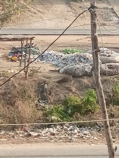 This image depicts an outdoor collection or recycling area within Ebiya, Nigeria. The scene is characterized by a dry, sandy terrain, partially covered with sparse, dry brown vegetation and some green shrubs. In the mid-ground, large, unsorted piles of plastic bottles are visible, predominantly clear or white, with a smaller accumulation of green bottles. Adjacent to these piles are several large, grey or black bags, appearing to contain sorted materials. To the left, a rudimentary wooden shelter with a corrugated roof stands over an accumulation of miscellaneous items. A wooden utility pole with multiple black wires strung across the frame occupies the right foreground. In the background, a paved road stretches horizontally, with a barren, light-brown dirt embankment rising behind it. No individuals, direct actions, or interactions are visible in the frame. Scattered debris, including various pieces of trash, is present across the ground.