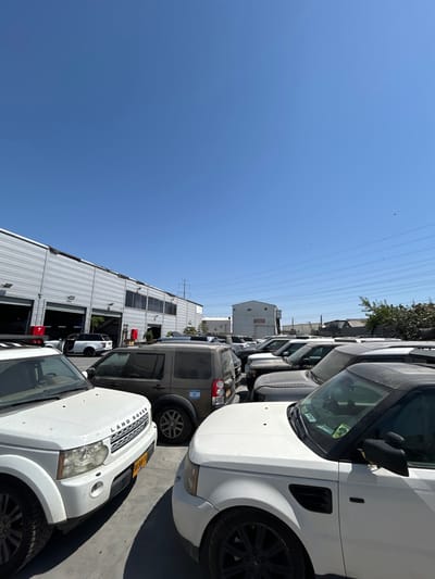 Rows of Land Rover SUVs fill a parking lot beside a large, white industrial building. The vehicles are a mix of white, gray, and tan colors. A clear blue sky is overhead, with power lines visible. The image is rotated to a 90-degree angle. One SUV in the center has a visible flag on the rear tire.