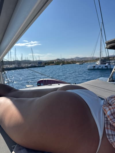 The image captures a sunlit nautical scene, viewed from a low perspective on board a vessel, in Lavreotiki Municipal Unit, Greece. In the immediate foreground, the lower back, buttocks, and upper thighs of a person are visible, lying prone on a grey, textured surface, likely a sunpad or deck cushion. The skin is tanned. The person is wearing white bikini bottoms, and a white, orange, and brown plaid garment is draped across their lower right side. To the left, a grey boat structure, possibly a canopy or roof edge, extends diagonally across the top left corner of the frame.

The midground features calm, deep blue water with numerous shimmering sun reflections creating circular bokeh effects. Masts and rigging from multiple sailboats are visible across the horizon line. A prominent white catamaran is anchored in the mid-right background, and other boats of various sizes are scattered across the water, some appearing to be docked in a distant marina.

In the background, a hilly coastline or distant landmass is visible under a clear, bright blue sky with sparse, wispy white clouds in the upper left quadrant. The scene is illuminated by strong natural sunlight, casting visible shadows on the person's left side and highlighting the water's surface.