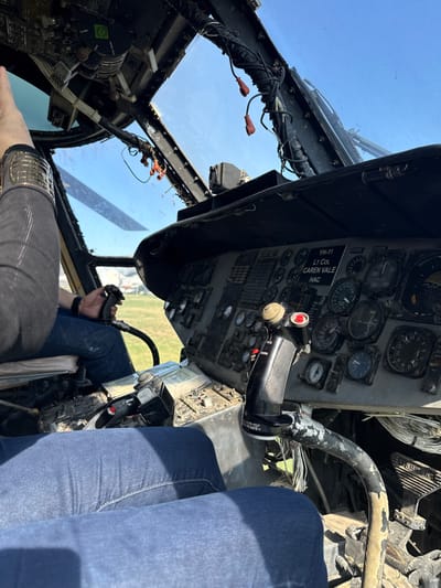 The interior of a helicopter cockpit shows various gauges and controls. A black control stick with a red button is prominently displayed. A plaque reads "YH-11 LT COL CAREN VALE HAC." One person wearing blue jeans occupies the passenger seat, while another person in gray pants sits in the pilot's seat with their hand on the controls. Outside, the sky is visible through the windshield, suggesting clear weather.