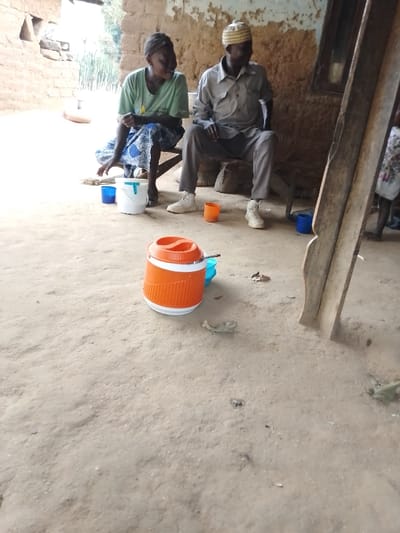 Two elderly individuals sit on wooden furniture near a mud wall structure. The woman wears a t-shirt and patterned skirt. The man has on a light-colored shirt, trousers, and cap. Various cups and a white bucket are on the ground, along with an orange water container. A wooden structure is visible to the right, and a third person stands partially in the background. The ground is dirt.