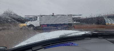 The image, captured from the interior of a vehicle through a rain-splattered windshield, depicts an outdoor industrial scene in Nazarbek, Uzbekistan. A white GAZelle-type flatbed truck with light-colored metal side rails occupies the mid-ground, loaded with several long, dark, rectangular metal beams or pipes extending over the cab. A single male individual, wearing a dark shirt, stands upright within the truck's bed, positioned near the front of the cargo. To the immediate left and partially obscured by the white truck's cab, a small, yellow utility vehicle is visible. The foreground shows the vehicle's dashboard and the lower portion of the windshield, covered with raindrops and a thin layer of what appears to be slush or dirt. The background features an extensive outdoor storage area, including numerous stacks of reddish-brown pipes arranged horizontally, and dark-colored industrial buildings with corrugated metal structures. A dark mound of earth or debris is present on the far left. The sky is overcast, and the ground appears wet and muddy.