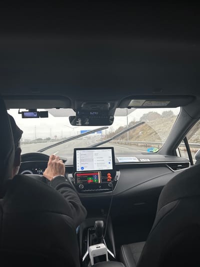 The image captures the interior of a car from the passenger's perspective, looking forward through the windshield. A driver, seen from the chest down, has their right hand on the steering wheel, wearing a silver bracelet and a dark long-sleeved garment. The steering wheel partially obscures a digital speed display showing "112", likely indicating 112 km/h.

The car's dashboard is equipped with multiple electronic devices. A large tablet, mounted centrally, displays what appears to be Google Maps, showing a list of options like "Ajustes" (Settings), "Contactos" (Contacts), "Lugares de interés" (Points of interest), "Trabajo" (Work), and "Casa" (Home). The time "19:23" is visible in the top right corner of the tablet screen. Below the tablet, a smartphone is horizontally mounted, displaying colorful app icons.

To the left, near the rearview mirror, a car radio or central unit shows "6:29" on its display. Above it, a sun visor is down, bearing "AIRBAG" text and warning symbols. A dashcam is mounted on the windshield, to the left of the rearview mirror. Charging cables are visible, connecting to a white portable charger or inverter located near the automatic gear shift.

Through the windshield, the view is of an overcast sky and a road with what looks like wintery conditions, possibly snow or frost on the distant ground. Wiper blades are visible in the foreground, suggesting recent or ongoing precipitation. The scene suggests the car is in motion on a highway or major road. Given the provided context, the location is Guadarrama, Spain.