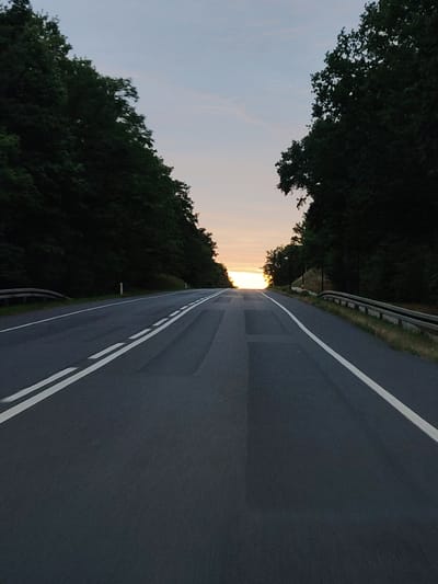 The image depicts a road in Rajszew, Poland, leading towards a sunset. The road is paved and has double white lines marking the lanes. On both sides of the road, dense, dark green trees line the path, creating a tunnel-like effect. The sky above is a gradient of pale blue and pink, with the setting sun a bright yellow orb visible at the horizon. The scene appears to be devoid of people or vehicles, suggesting a quiet and serene moment.