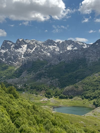 An eye-level view of a mountain landscape in Brskut, Montenegro. A tall, rocky mountain with patches of snow stands adjacent to a green, forested slope that leads down to a small lake. A cluster of buildings, likely residences or tourist accommodations, are visible near the base of the slope. Above the mountain is a blue sky with scattered white clouds.