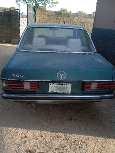 A teal Mercedes-Benz 200, seen from the rear, exhibits a weathered appearance. Its rear window is dusty, and a chain drapes along the edge. The license plate reads "KOH-50ES" and indicates origin from Adamawa State, Nigeria. The car is parked on dirt ground, with a brick wall and gate visible in the background. The car's chrome bumper shows signs of age, while the Mercedes-Benz emblem is prominently displayed on the trunk.