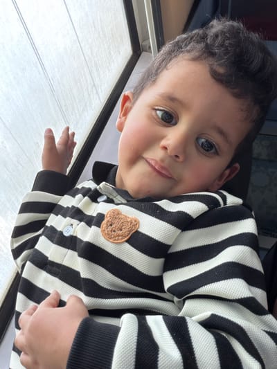 A young child with curly dark hair and brown eyes looks up at the camera. They are wearing a black and white striped shirt with a brown teddy bear patch. The child is next to a window with a white blind. One hand rests on the blind. The image is rotated to the left. The child's expression is neutral, with a slight hint of a smile.
