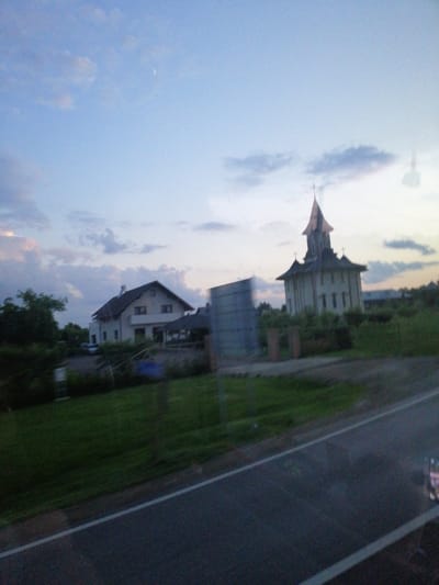 The image captures a rural scene at dusk in Dărmănești, Romania. On the left, a modern house with a dark roof and light-colored walls is visible amidst trees and a green lawn. To the right, a church with a distinctive pointed roof, reminiscent of traditional Romanian architecture, stands against a pale sky. The church is surrounded by greenery, and a large, grey, rectangular structure is situated to its left. A paved road with a white line runs across the foreground, suggesting the perspective is from a vehicle moving along the road. The sky is a mix of blue and soft clouds, with a faint hint of the moon. The overall impression is one of a tranquil evening in a rural setting.