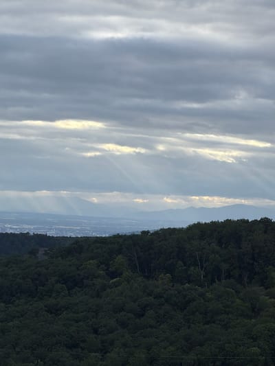 The image depicts a wide, elevated view of a landscape under a cloudy sky. In the foreground, a dense, dark green forest covers a hillside. Beyond the forest, a hazy cityscape stretches across a valley, with distant mountains forming a backdrop. Sunlight pierces through breaks in the clouds, creating visible rays that illuminate parts of the sky and the landscape below. The scene is devoid of any human presence or specific landmarks that would indicate a precise location within Sissach, Switzerland. The overall impression is one of a natural landscape with a distant urban area, under atmospheric lighting conditions.