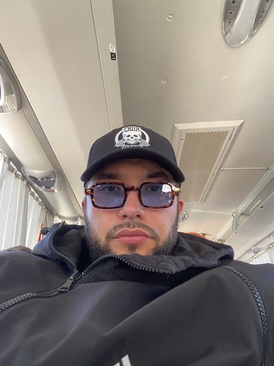 A low-angle, close-up, selfie-style photograph shows a man wearing a black trucker hat with a white skull logo and a black puffer jacket. He is also wearing brown-rimmed sunglasses. The man has a beard and appears to be in a vehicle, possibly a bus or train, with ceiling lights and air vents visible in the background. The interior of the vehicle is light-colored, with white curtains visible to the left. The image was taken in the city of Kaykino, Russia.