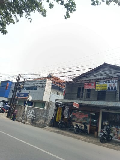 A street scene features a BRI bank building and adjacent two-story building with real estate advertisements hanging from its balcony. Several motorcycles are parked in front of a small storefront with a "WARTEC" sign. Graffiti adorns the concrete wall. Power lines crisscross above the buildings under a bright sky, partially obscured by tree branches. The image is taken from a slightly low angle from the street.