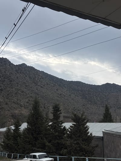 This image depicts a mountainous landscape in Lehvaz, Armenia, under an overcast sky. The upper portion of the frame shows a dark structural overhang and multiple overhead utility lines traversing horizontally, with a vertical support structure visible on the left. Below these lines, a prominent, dark, rocky mountain range with sparse vegetation dominates the background. In the mid-ground, a line of dark green conifer trees (likely junipers) stands in front of several low-rise buildings. One building's corrugated metal roof is visible behind the trees, with a small, multi-colored object (red, white, and blue) partially discernible on its surface towards the right. Another building with dark, possibly stone or brick, exterior walls is partially visible on the far right. In the lower foreground, a blue metal fence with vertical posts and a horizontal top rail runs across the scene. A white, older model sedan, consistent with a VAZ-2101 or similar Lada, is parked immediately behind this fence, facing slightly left. No individuals are visible.
