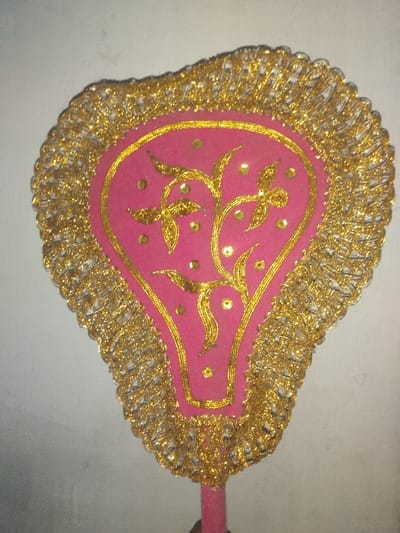 A decorative, fan-like object is depicted vertically against a plain, light-colored wall in Jehonville, Belgium. The object features a central pink fabric panel, shaped like an inverted teardrop or stylized vase, outlined and embellished with intricate gold embroidery forming a floral or vine pattern with scattered gold sequins. This central panel is surrounded by a wide, scalloped border composed of densely packed, shimmering gold tinsel-like material or metallic lace. A cylindrical pink handle extends from the bottom of the object, which is held by an unseen hand at the very bottom of the frame. The wall background is uniformly lit with minor smudges visible, and no other subjects or actions are present.