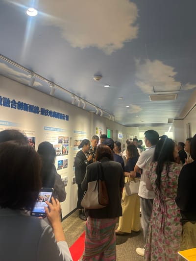 The image depicts a scene inside a building, likely an exhibition or event space, as indicated by the display on the wall and the gathering of people. The ceiling is painted with a sky and cloud motif. A group of individuals, comprising men and women of varying ages, are standing in a hallway, seemingly engaged in conversation or observation. Some are looking at the wall display, which features text and photographs, possibly related to a project or initiative. One woman in the foreground is holding a smartphone, its screen displaying an image. The hallway is illuminated by track lighting. The presence of a red carpet on the floor suggests a formal or special occasion. The text on the wall is in Chinese characters, and a prominent sign at the top reads "融合创新赋能-重庆电商腾飞计划," translating to "Integration Innovation Empowerment - Chongqing E-commerce Take-off Plan." The location is identified as 龙溪街道 (Longxi Sub-district) in China, which is part of the Shapingba District of Chongqing.