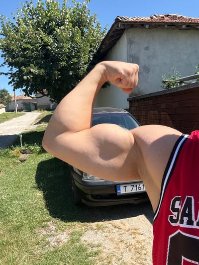 The image depicts a close-up of a man's flexed bicep, with his fist clenched and held aloft. He is wearing a red athletic jersey with white trim. Behind him, a black car is parked, with a license plate visible that reads "BG T 7161". The scene appears to be outdoors, with a rural setting evident. A dirt road and green grass are present, along with a house with a tiled roof and a large tree. The lighting suggests a sunny day. The car and surrounding buildings suggest a location in a village or town, possibly Konop, Bulgaria, given the license plate format.